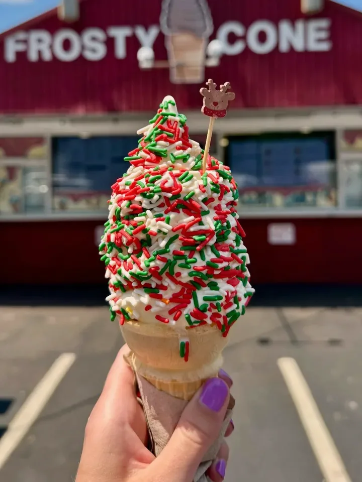 Vanilla soft serve cone covered in red, green and white sprinkles with a reindeer pick, held up in front of the Frosty Cone stand